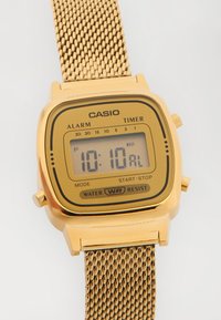 Relógio digital Casio em tom dourado com bracelete de malha, mostrando a hora 10:10, com funções de alarme e cronômetro, e etiqueta de resistência à água.
