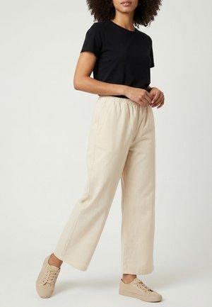 Femme portant un T-shirt noir à manches courtes, un pantalon large beige avec une taille élastique et des baskets beige à lacets, debout devant un fond uni.