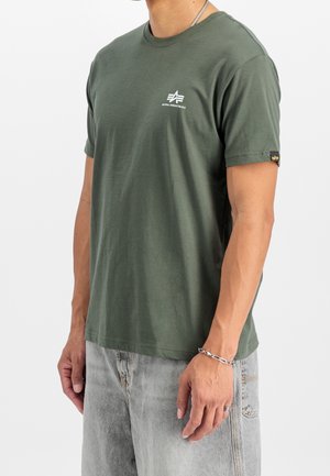Mężczyzna w oliwkowej koszulce Alpha Industries z małym białym logo na piersi i wyblakłych szarych dżinsach, stojący na jednolitym białym tle.