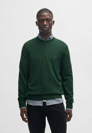 Groene gebreide trui met een ronde hals, reguliere pasvorm en geribde manchetten. Voorzien van een logopatch op de borst en gelaagd met een lichtgrijze shirt eronder.