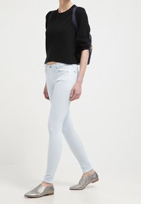 Suéter negro corto, jeans ajustados azul claro y zapatos planos plateados. La modelo lleva una mochila azul oscuro, de pie sobre un fondo liso.