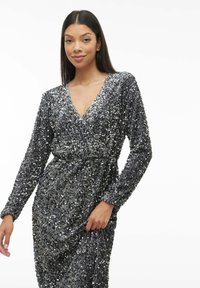 Abito avvolgente argento in paillettes con maniche lunghe e scollatura a V, presenta una vita arricciata e una gonna fluente, creando un aspetto testurizzato e luccicante.