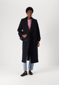 Double-breasted navy coat with notched lapels, worn over a light pink top, paired with light blue jeans and black loafers.