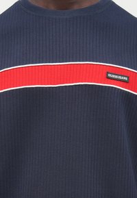 Tumesinine soonikkoest ümarkaelusega dressipluus punase horisontaalse triibu ja valge aktsendiga. Varustatud musta "GUESS JEANS" sildiga.