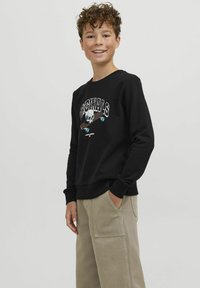 Felpa nera con grafica di teschio e skateboard. Polsini e bordo a coste, indossata con pantaloni cargo beige. Texture liscia, design casual.