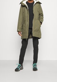Olive green parka with a faux fur lined hood, zipper front, and two side pockets. Black pants and sneakers with teal accents complete the look.