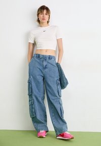 Vit kortärmad t-shirt med svart logga, blå denim cargo byxor med sidofickor, och rosa sneakers med vita detaljer. Enfärgad och avslappnad passform.