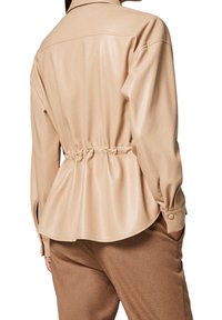 Chemise beige, brillante, avec une coupe décontractée, taille cintrée, manches longues et détails de boutons. Présente des fentes latérales à l'ourlet.