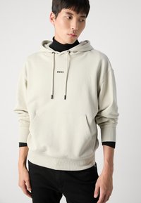 Sudadera con capucha color beige claro con detalles en negro, gran bolsillo frontal y logo 'BOSS' en negro en el pecho. Material de algodón.