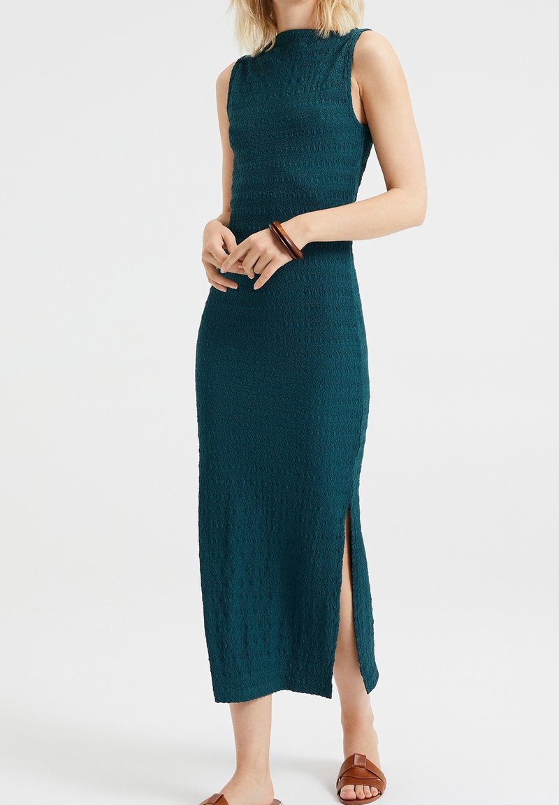 Ärmlös turkos texturerad midiklänning med sidoslits, bärs med brunt träarmband och bruna platta sandaler på ljus bakgrund.