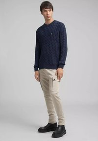 Marineblauwe kabelgebreide trui met lange mouwen, gecombineerd met lichtbeige cargobroek met zijzakken en zwarte enkellaarsjes.