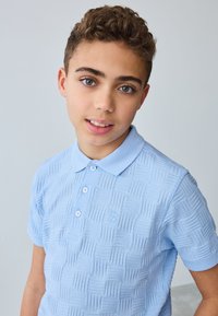 Vaaleansininen pikeepaita, jossa on teksturoitu kudottu kuvio, lyhyet hihat ja kaksi nappia. Rinnassa brodeerattu logo.