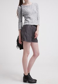 Grau strukturiertes Oberteil mit langen Ärmeln, schwarze hochgeschnittene Shorts und schwarze ankle boots mit silbernen Nieten. Das Model hält eine schwarze Handtasche mit Fransen.
