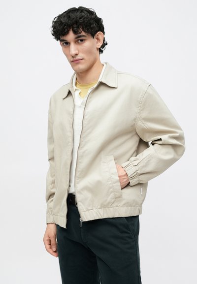 Bežinė lengva bomber tipo striukė su priekiniais užtrauktukais, šoniniais kišenėmis ir elastingomis rankovėmis, dėvima ant baltos ir geltonos sluoksniuotos marškinės.