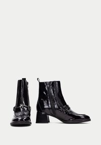 Botines de charol negro con tacón ancho, que presentan una cremallera lateral, un hebilla decorativa y una textura suave y brillante.