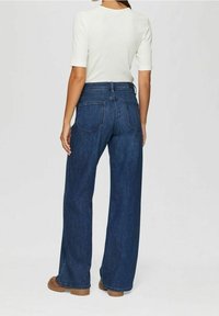 Mujer vestida con jeans oscuros de pierna ancha y cintura alta, camiseta blanca de manga corta y zapatos marrones, de pie de espaldas frente a un fondo liso.