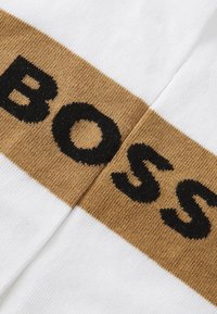 Λευκό ύφασμα με οριζόντια καφέ λωρίδα με μαύρο κείμενο "BOSS". Το υλικό φαίνεται πλεγμένο με λεία υφή.