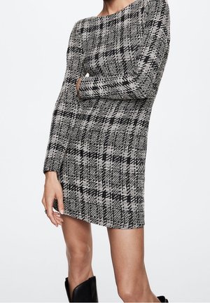 Femme portant une mini-robe à manches longues pied-de-poule noir et blanc, bras croisés et bottes noires sur fond blanc.