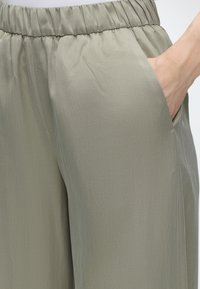Pantalon vert olive clair à taille élastique avec une main partiellement dans la poche latérale droite, montrant une texture de tissu lisse.