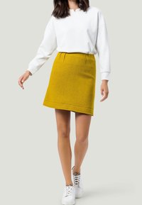 Gonna di lana gialla con una leggera forma a A, caratterizzata da pieghe frontali e un orlo che si trova sopra il ginocchio, abbinata a un maglione bianco.