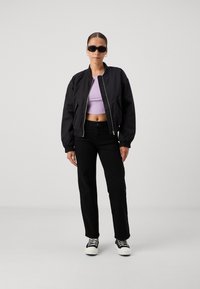 Black bomber jacket over a lavender crop top, black straight-leg pants, and black and white sneakers. Accessorized with dark sunglasses.