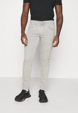Man draagt lichtgrijze slim-fit jeans, zwart t-shirt, witte sokken en zwarte sneakers, terwijl hij staat tegen een effen lichte achtergrond.