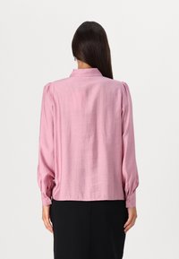 Blouse à manches longues en tissu rose clair, avec un col et des détails de poignet boutonnés. Texture lisse avec une coupe légèrement ample.