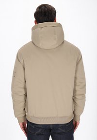 Beige hættejakke med quiltet tekstur, ribbede manchetter og kant, samt en lomme på venstre ærme. Bagfra, med minimale detaljer.