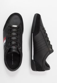 Le sneakers nere presentano una tomaia in pelle liscia, dettagli in suede, sei occhielli per i lacci e una suola bianca con un design testurizzato. Strisce rosse e bianche sui lati.