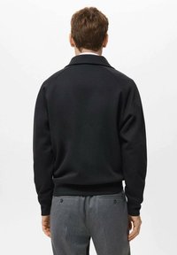 Sudadera negra con un diseño sencillo, puños y dobladillo acanalados. Con cuello clásico. Combina con pantalones grises. Vista trasera.