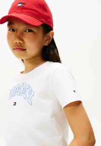 Hvid bomulds t-shirt med blå "TOMMY" tekst, rund hals, korte ærmer og lille logo lap i rød, hvid og blå på ærmet. Rød cap.