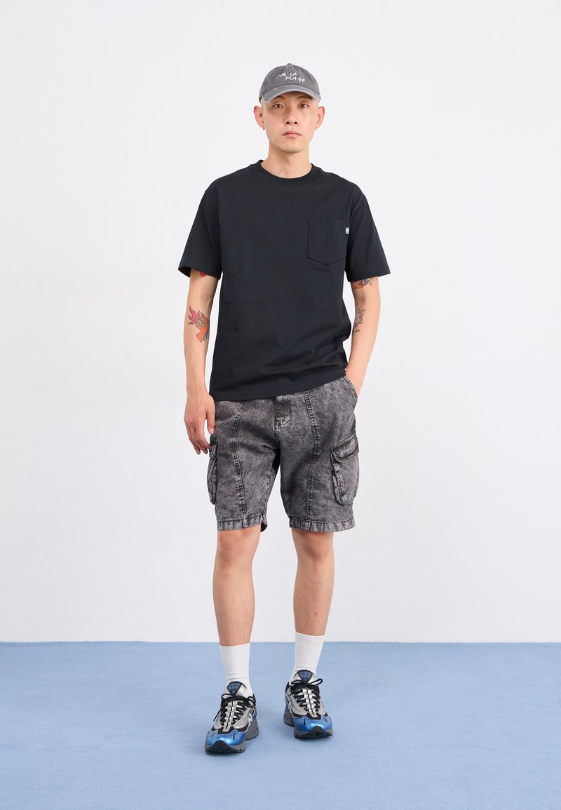 Mann trägt ein schwarzes T-Shirt mit Taschen, graue Cargo-Shorts, blaue Sneaker, weiße Socken und eine graue Mütze und steht auf einem blauen Boden vor einem weißen Hintergrund.