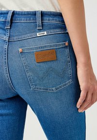 Blå denimjeans med en brun läderlapp på bakfickan som har ett sydd zick-zack-mönster och kopparnitar. Blekt textur.