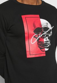 Felpa nera con una grande grafica di teschio argentato su uno sfondo rettangolare rosso, con sovrapposizione di testo bianco. Morbida texture del tessuto.
