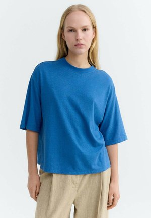 Junge Person trägt ein lockeres blaues Kurzarmhemd und beige Hose, steht vor einem schlichten weißen Hintergrund.