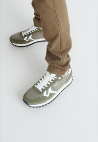 Zapatillas deportivas en verde y gris con cordones y acentos blancos, que presentan una textura de ante y malla, sobre una suela de goma negra.