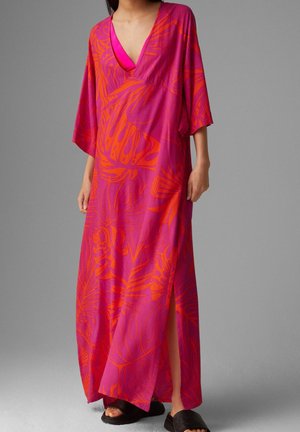 Vrouw draagt een losse, lange roze jurk met oranje tropische bladpatronen, een diepe V-hals en een zijsplit, gecombineerd met zwarte slippers.