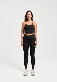 Zwarte sportbeha met dunne banden en logo. Gemaakt in combinatie met hoog-getailleerde zwarte leggings met een crossover-ontwerp. Grijze sneakers.