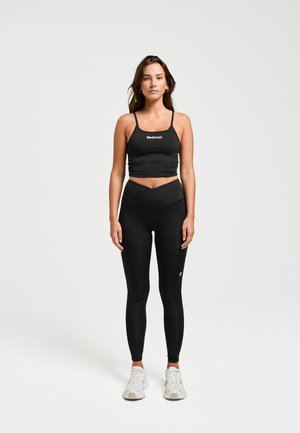 Schwarzer Sport-BH mit dünnen Trägern und Logo. Kombiniert mit hochgeschnittenen schwarzen Leggings mit Überkreuzdesign. Graue Sneaker.