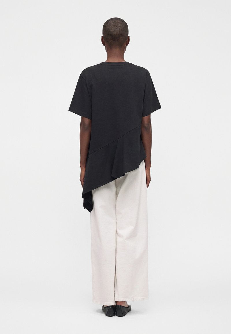 Person, die mit dem Rücken zur Kamera steht, trägt ein schwarzes asymmetrisches Kurzarmtop, weiße Hosen mit weitem Bein und schwarze Schuhe vor einem einfarbigen Hintergrund.