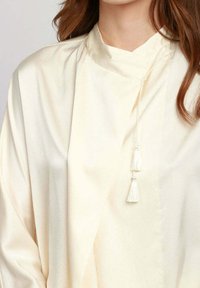 Top color crema con colletto alto, caratterizzato da una texture liscia e lucida e accenti a nappina. Il design è oversize e dal taglio ampio.