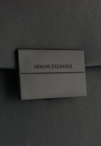 Fekete bőr táska sima textúrával, fekete téglalap alakú fémcímkével, amelyen az "ARMANI EXCHANGE" felirat található.