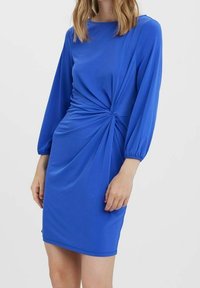 En kvinna visar upp en åtsittande, klarblå klänning med långa ärmar och en vridning vid midjan, mot en enkel vit bakgrund.