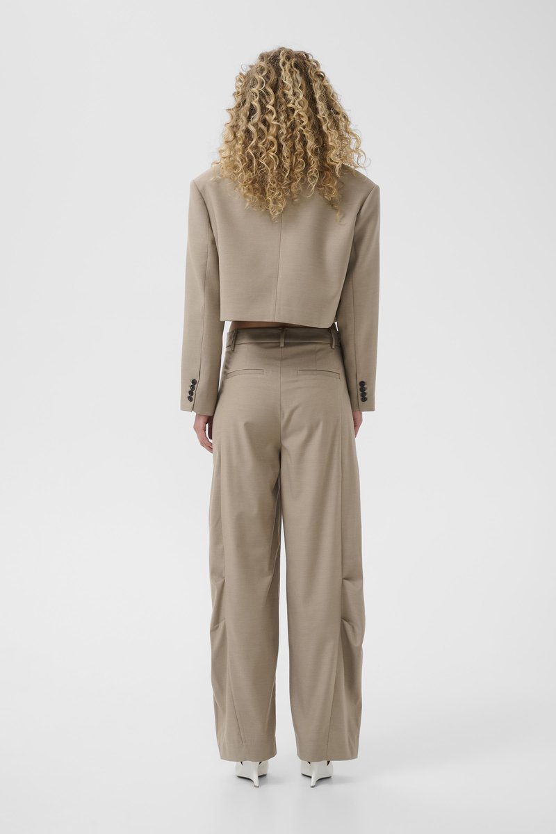 Woman with curly blonde hair stands facing away, wearing a beige cropped blazer, matching wide-leg trousers, and white pointed shoes.