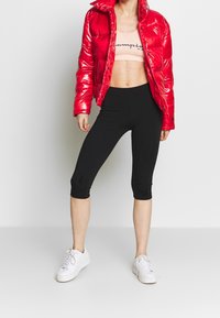 Czerwona, błyszcząca kurtka puchowa z wysokim kołnierzem, noszona na jasnoróżowym krótkim topie z czarnymi napisami, do tego czarne legginsy capri i białe trampki.