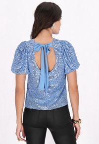 Top blu in paillettes con maniche a sbuffo, schiena scoperta e chiusura con fiocco azzurro. Presenta un tessuto lucido e testurizzato e un design aderente.