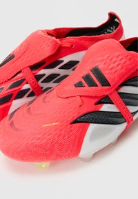 Φωτεινά κόκκινα ποδοσφαιρικά παπούτσια Adidas με μαύρες και λευκές λεπτομέρειες, καλυμμένο μπροστινό μέρος και καρφιά στη σόλα για πρόσφυση.