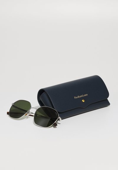 Óculos de sol com lentes verdes e armação prateada, repousando ao lado de uma caixa escura azul-marinho com um logo dourado e fecho de pressão.