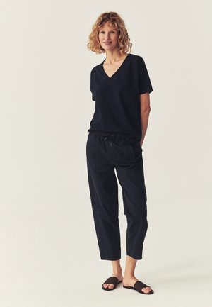 Frau mit lockigem blondem Haar, die ein schwarzes V-Ausschnitt Kurzarmshirt, schwarze Hose mit Kordelzug und schwarze Slip-On-Sandalen trägt, steht mit den Händen hinter dem Rücken.