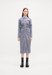 Abito a camicia a quadri blu e bianco, con bottoni sul davanti, maniche lunghe, vita arricciata, lunghezza al ginocchio, abbinato a stivali neri alla caviglia.